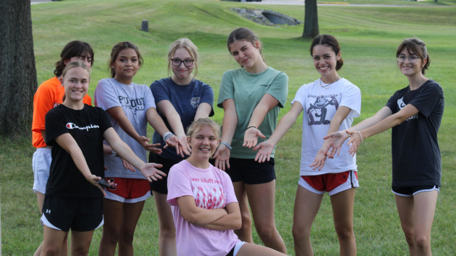 Girls cross country team jiggles jazz hands towards Olivia Gibson to pose for photographer pre-workout. 