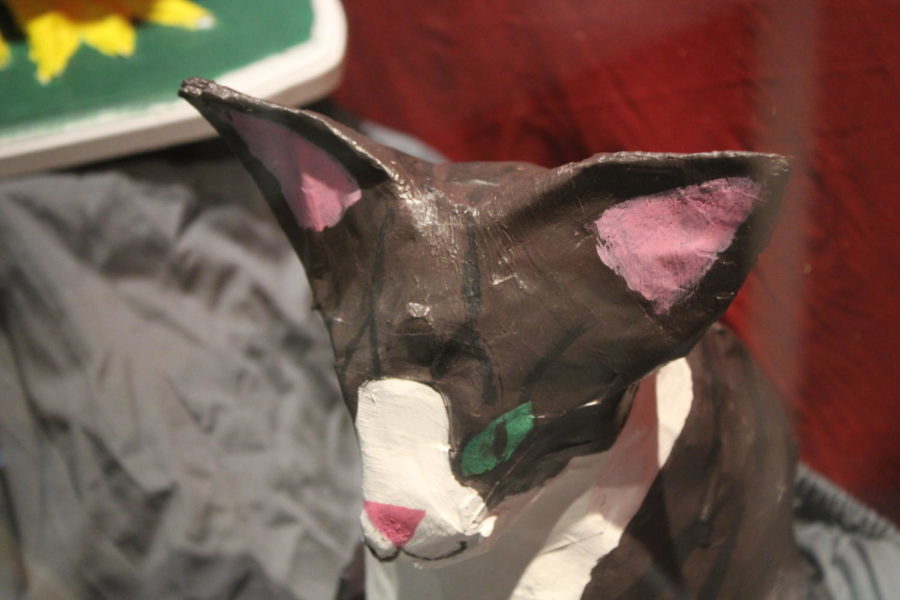 This Papier-mâché cat rests in a case on the bridge between the English hallway and the art gallery. The cat has white and mocha-colored fur, along with green eyes. It rests not by itself, but with other similar pieces, including a light brown deer with huge antlers, a white rat with red eyes, and a green flamingo. 