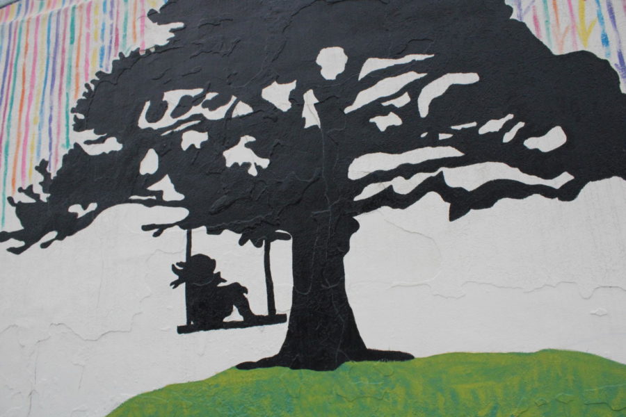 A mural depicting a small child taking cover from the rain under a tree in the Rainy Play mural found across the west side of Bonus Pints and Record Farm.