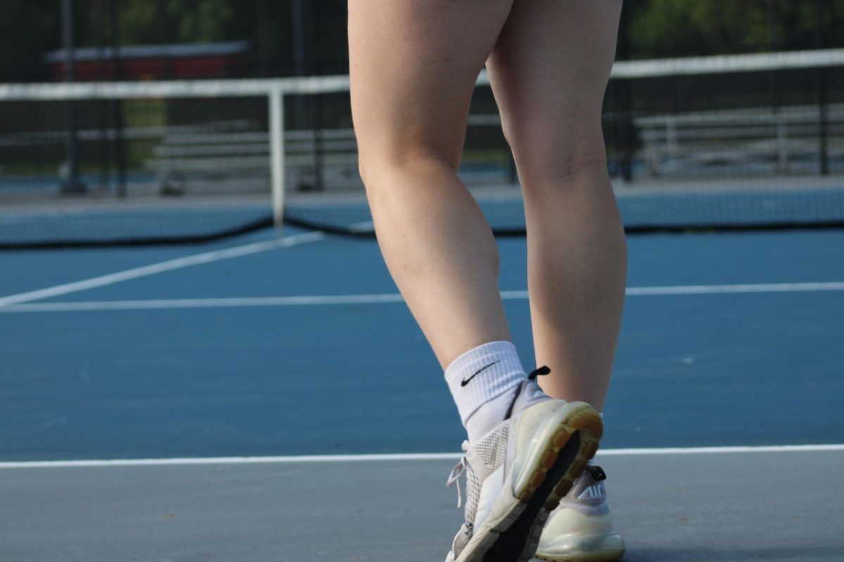 Sophomore tennis varsity player stands behind the white line. She looks off into the distance as she is motioned where to stand. 