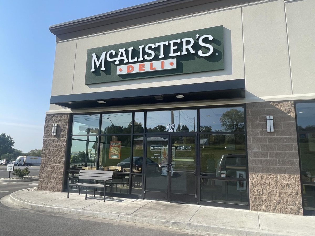 McAlister's Deli entrance reflects the blue sky and its surroundings. Inside the deli, several people are lining up to get their order.