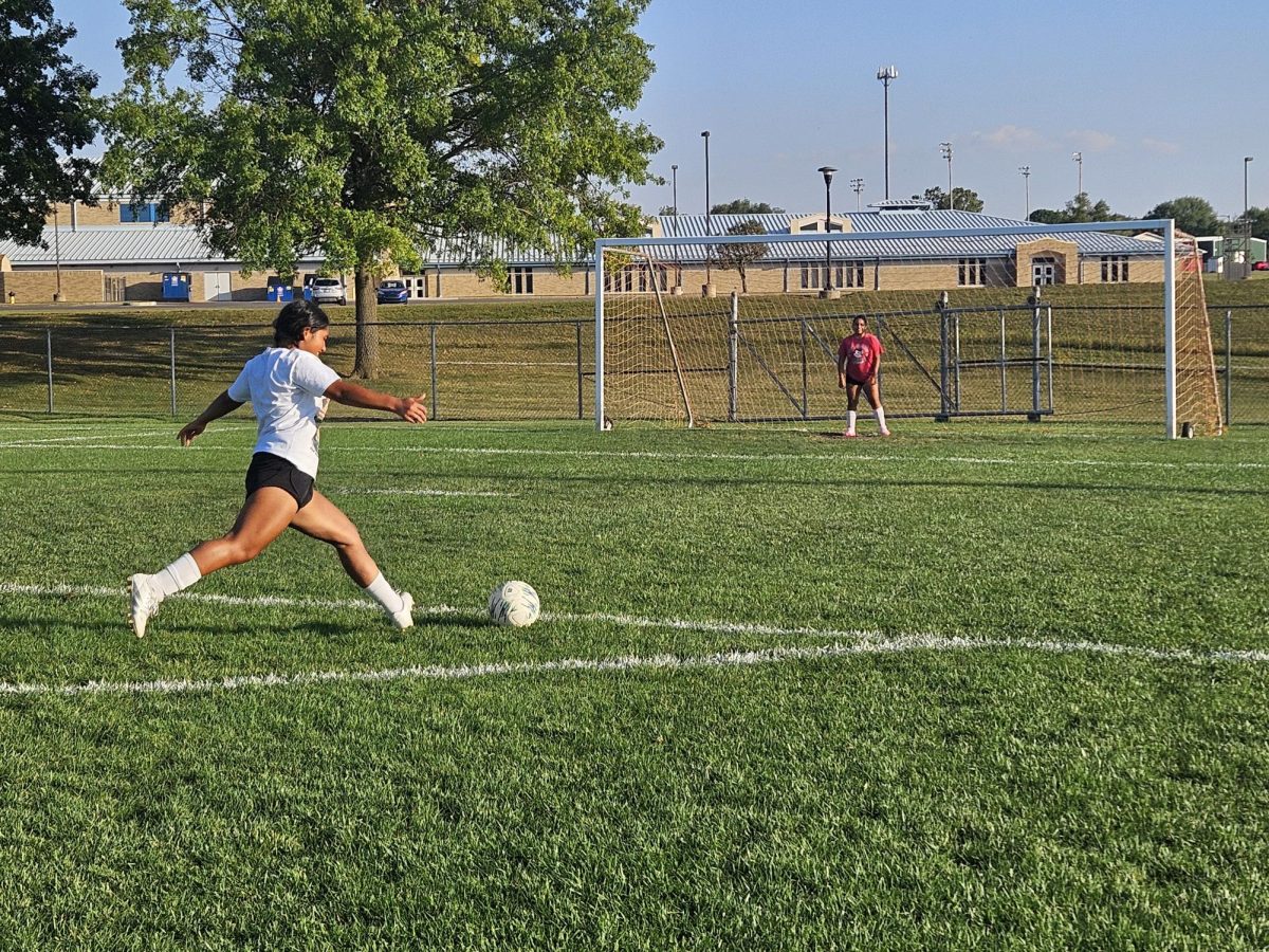 Acting like a goalkeeper, Yesenia Pascual-Sebastian stands on the goal line as Angelica Hernandez steps in hard to shoot the ball. Before soccer practice starts, players freestyle, pass, or shoot as their personal warm-up.